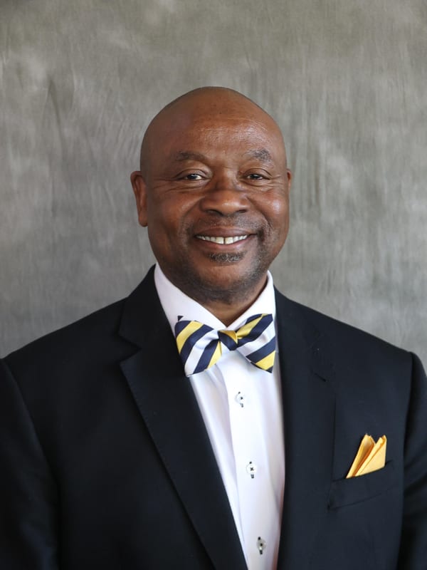 Bruce J. Young smiling, in a navy suit with a navy and gold striped bow tie and a gold pocket square